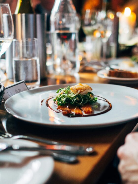 Close-up of a plated dish with greens and sauce on a white plate at a restaurant table, with a bread basket and wine glasses nearby.