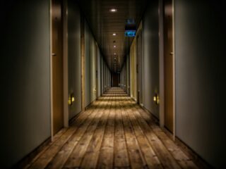 Long, dim hotel hallway with doors on both sides and warm overhead lights, extending toward a distant exit sign.