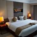 Hotel bedroom with a tufted headboard wall, built-in fireplace, two lamps, and two white beds with brown runners and pillows.