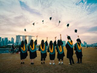 Graduates in black gowns with yellow sashes toss their caps in the air at sunset, with a city skyline in the background.