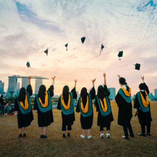 Graduates in black gowns with yellow sashes toss their caps in the air at sunset, with a city skyline in the background.