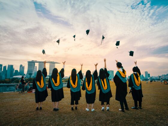 Graduates in black gowns with yellow sashes toss their caps in the air at sunset, with a city skyline in the background.