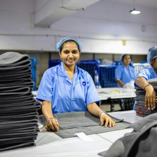 Two women in blue uniforms and hairnets fold dark fabric at a textile production line, with stacked sheets in the foreground.