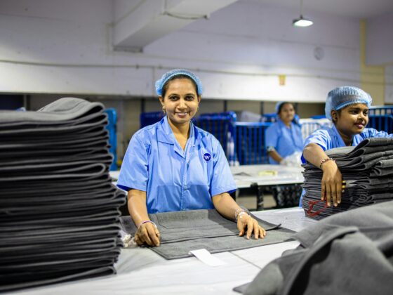 Two women in blue uniforms and hairnets fold dark fabric at a textile production line, with stacked sheets in the foreground.