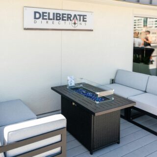 Outdoor patio seating area with four gray-cushioned chairs around a dark wicker table topped with a glass insert and blue decorative stones on a light deck.
