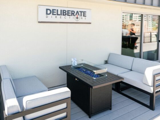 Outdoor patio seating area with four gray-cushioned chairs around a dark wicker table topped with a glass insert and blue decorative stones on a light deck.