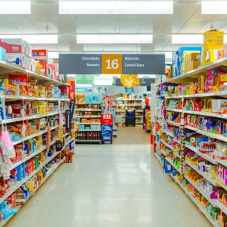 A brightly lit supermarket aisle filled with colorful packaged snacks on shelves, with a central sign listing Chocolate Sweets, 16, Biscuits and Cereal bars.