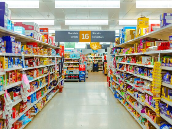 A brightly lit supermarket aisle filled with colorful packaged snacks on shelves, with a central sign listing Chocolate Sweets, 16, Biscuits and Cereal bars.
