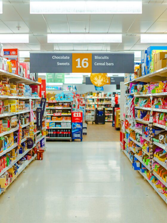 A brightly lit supermarket aisle filled with colorful packaged snacks on shelves, with a central sign listing Chocolate Sweets, 16, Biscuits and Cereal bars.