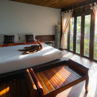 A tropical-style hotel bedroom with a large wooden bed, white linens, orange runner, and decorative pillows facing glass doors.