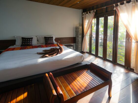 A tropical-style hotel bedroom with a large wooden bed, white linens, orange runner, and decorative pillows facing glass doors.