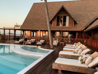 Resort pool area with wooden deck, lounge chairs, and a thatched-roof building; a person stands by the pool at sunset nearby palm trees.