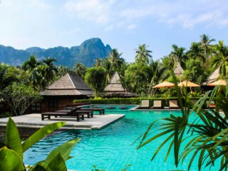 Poolside tropical resort with turquoise water, thatched villas, palm trees, and mountain backdrop.