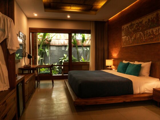 Hotel room with a large wooden bed, blue bedding and teal pillows, warm lamps, and a garden view through glass doors.