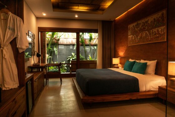 Hotel room with a large bed, teal pillows, warm lighting, and wooden furnishings opening to a lush garden through a glass door.