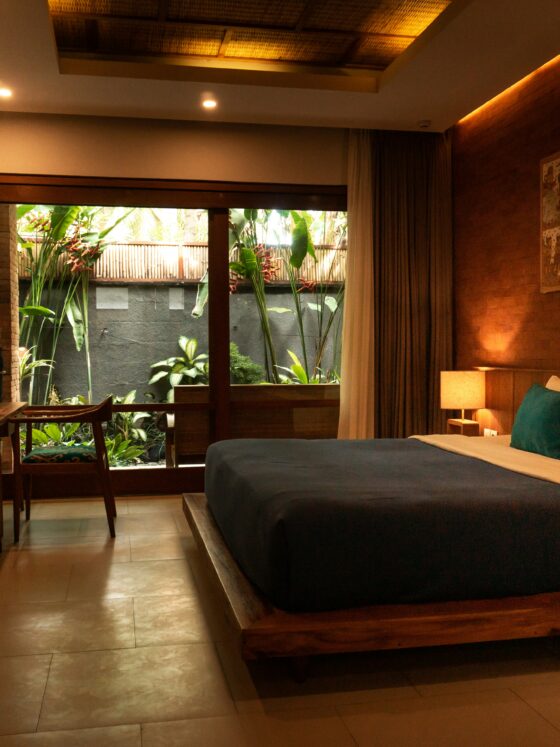 Hotel room with a large bed, teal pillows, warm lighting, and wooden furnishings opening to a lush garden through a glass door.