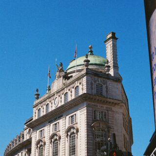 Facade of a historic London building with a green copper dome, statues, flags, and arched windows against a bright blue sky.",