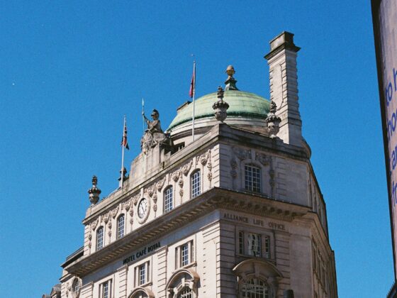 Facade of a historic London building with a green copper dome, statues, flags, and arched windows against a bright blue sky.",