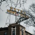 Brick hotel building with a yellow vertical sign reading 'Hotel' and a flag, framed by bare tree branches overhead in gray sky.