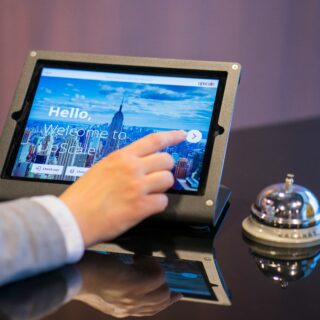 Person taps a tablet kiosk showing a cityscape welcome screen with the text “Hello, Welcome to Upscale.”