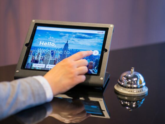 Person taps a tablet kiosk showing a cityscape welcome screen with the text “Hello, Welcome to Upscale.”