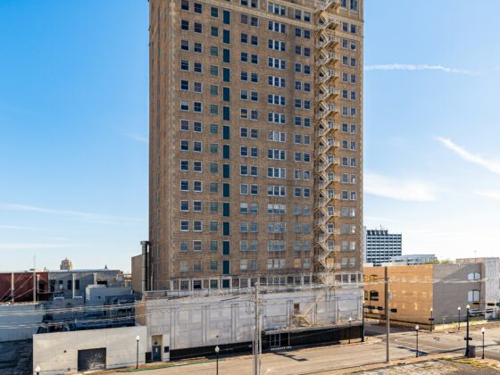 Tall brick office building with external fire escape along the right side and satellite dishes on the roof against a clear blue sky.