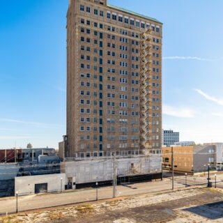 Tall beige brick high-rise with external fire escape on the right and antennas on the roof against a clear blue sky, urban street foreground.