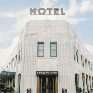 Art deco hotel exterior with a large lit 'HOTEL' sign atop a white stone building; entrance doors below.