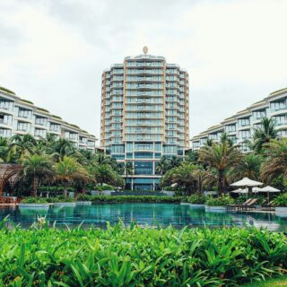 Resort complex with a tall central tower, two low-rise wings, and a large swimming pool surrounded by palm trees in the foreground.