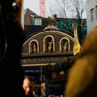 Crowded city street at a Christmas market; a decorative storefront with arched windows and statues, reading 'HEINZELS WINTERMÄR' with a red crane behind.