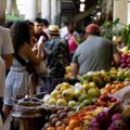 Indoor market scene with fruit stalls and shoppers; baskets of mangoes, dragon fruit, and oranges in the foreground.