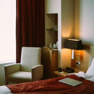 Hotel room with burgundy bed runner, beige chair, and wooden desk with lamp and gadgets near the bed.