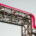 Exposed steel framework supporting a bright pink curved pipe against a pale sky backdrop.