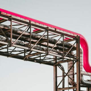 Exposed steel framework supporting a bright pink curved pipe against a pale sky backdrop.