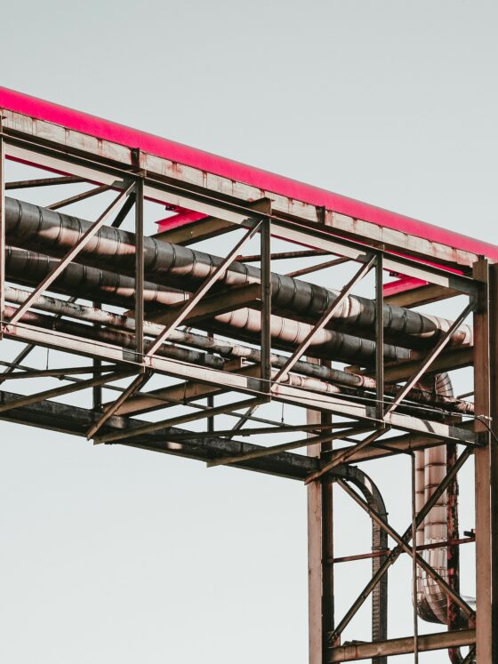 Exposed steel framework supporting a bright pink curved pipe against a pale sky backdrop.