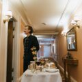 Waiter in a dark formal uniform stands in a peach-colored hallway beside a table set for a formal dinner with flowers and glassware in the foreground.