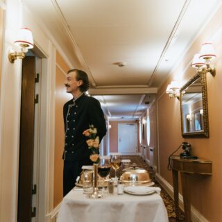 Waiter in a dark formal uniform stands in a peach-colored hallway beside a table set for a formal dinner with flowers and glassware in the foreground.
