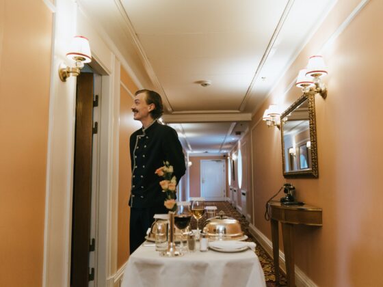 Waiter in a dark formal uniform stands in a peach-colored hallway beside a table set for a formal dinner with flowers and glassware in the foreground.