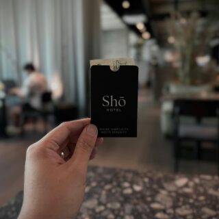 Close-up of a hand holding a black Shō Hotel card with a small paper insert at the top, in a hotel lobby. The background is blurred.