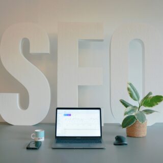Large white 3D letters spell 'SELF' on a desk with a laptop, mug, phone, and plant nearby.