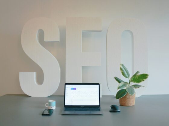 Large white 3D letters spell 'SELF' on a desk with a laptop, mug, phone, and plant nearby.