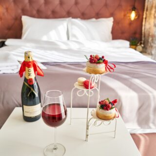 Champagne bottle and wine glass on a white table with a berry-topped pastry stand in front of a bed with white pillows and a pink tufted headboard in a cozy room.