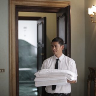 Man in a white shirt and black tie holding a stack of folded towels in a doorway of a vintage, warmly lit room.