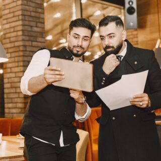 Two men in smart black outfits study a tablet and papers together in a warmly lit restaurant cafe setting.”,