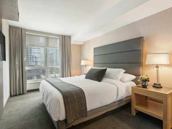 Hotel room with a large upholstered headboard, bed, two nightstands with lamps, and a city view through the window.