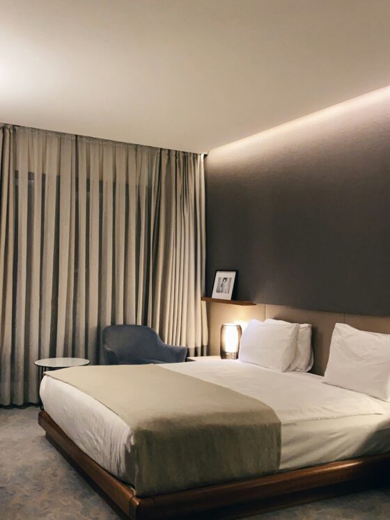 Hotel bedroom with a king bed, beige linens, gray accent wall, sheer curtains, and a nightstand lamp with a framed photo nearby on the shelf.