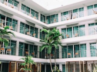 White multi-story hotel/condo building with glass-paneled balconies and wooden chairs along a courtyard, framed by palm trees.