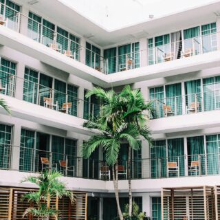 White multi-story hotel/condo building with glass-paneled balconies and wooden chairs along a courtyard, framed by palm trees.
