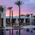 Poolside at a modern white resort during sunset, with tall palm trees and lounge chairs lining the pool edge.