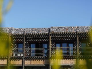 Stone building with a wooden pergola and balcony, clear blue sky behind, and greenery in the foreground with a white umbrella at the bottom blends in.